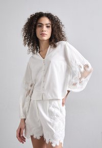 Top blanco de lino con escote en V, botones frontales y mangas voluminosas que presentan un intrincado detalle de encaje. Combinado con shorts blancos bordados.