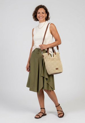 Frau trägt ein ärmelloses weißes Top, einen olivgrünen Rock mit Gürtel, braune Sandalen und hält eine beige geflochtene Schultertasche, lächelt in die Kamera.