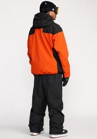 Veste de neige orange et noire avec capuche, présentant une coupe ample et des panneaux contrastants. Porte avec un pantalon de neige noir et des gants.