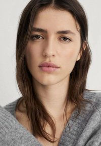Modella che indossa un morbido pullover grigio chiaro con scollatura a V. Capelli castano scuro e lunghi, che incorniciano il viso su uno sfondo neutro.