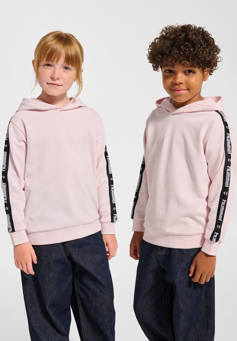 Lichtroze hoodies met zwarte zijkanten voorzien van logo-tekst, gecombineerd met losse donkere denimspijkerbroeken. Zacht, gestructureerd materiaal, casual pasvorm.