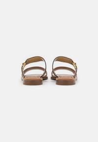 Sandali in pelle marrone con design senza retro, sottili cinturini e un accento in hardware dorato, poggiati su una suola piatta marrone testurizzata.