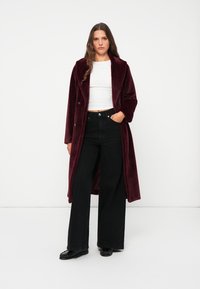 Burgundy long coat made from soft fabric, featuring a notched collar and two buttons, paired with black wide-leg pants and black shoes.