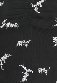 Černý žebrovaný top s bílými květinovými vzory, se shromážděním uprostřed, které zdůrazňuje strukturu a design.