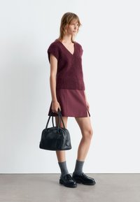 Burgunderfarbener Strickpullover mit V-Ausschnitt, kombiniert mit einem burgunderfarbenen Minirock. Das Model hält eine schwarze Lederhandtasche und trägt graue Socken und schwarze Schuhe.