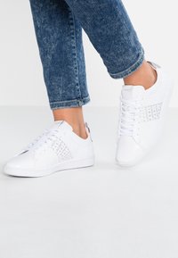 Zapatillas blancas con costados texturizados, cordones y un logo sutil. Material suave, punta redondeada y suela acolchada. Llevadas con vaqueros azules cortos.