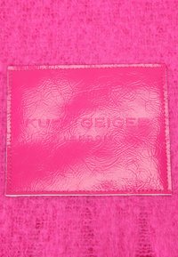 Sfondo in feltro testurizzato fucsia pink con un'etichetta in pelle lucida rosa, impressa con "KURT GEIGER LONDON" al centro.