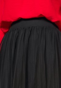 Gonna in tulle nera con una ampia vita elastica. Il tessuto presenta pieghe verticali, trame sovrapposte e una finitura traslucida. Abbinata a una maglietta rossa.