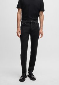 Black denim jeans with a slim-fit design, five-pocket style, and a subtle brand tag. Worn with a black short-sleeve shirt and black shoes.