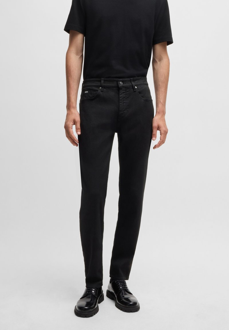Black denim jeans with a slim-fit design, five-pocket style, and a subtle brand tag. Worn with a black short-sleeve shirt and black shoes.