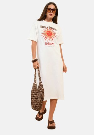 Žena nosiaca biele midi tričkové šaty s oranžovou slnečnou grafikou a textom, hnedé slnečné okuliare, masívne sandále, náramky a nesie tkanú tašku.