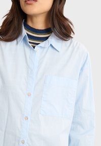 Camicia button-up azzurro chiaro con taschino sul petto, design con colletto e bottoni perlati. Indossata sopra un dolcevita aderente a righe.