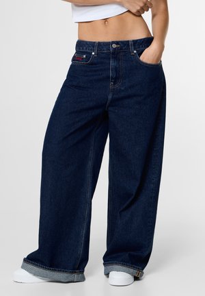 Persoon die donkerblauwe wijde jeans draagt met omgeslagen zomen en witte sneakers, bij de taille bijgesneden, waarbij een deel van de buik en een hand in de zak zichtbaar is.