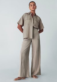 Béžový dvoudílný outfit skládající se z krátkého rukávu košile s kapsami a kalhot s širokými nohavicemi, vyrobený z lehké, strukturované tkaniny.