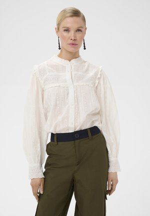 Femme aux cheveux blonds attachés, portant un chemisier en dentelle blanche avec des boutons et un pantalon vert olive avec une ceinture noire, debout sur un fond blanc.