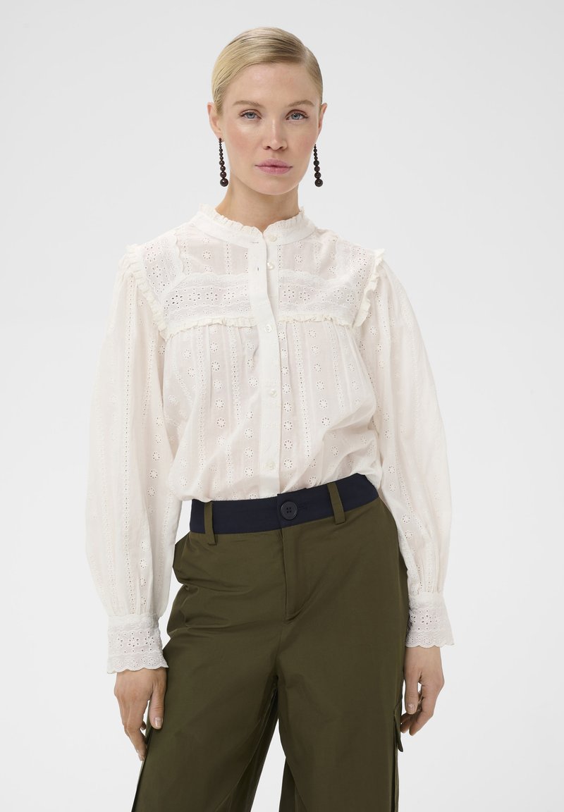 Femme aux cheveux blonds attachés, portant un chemisier en dentelle blanche avec des boutons et un pantalon vert olive avec une ceinture noire, debout sur un fond blanc.