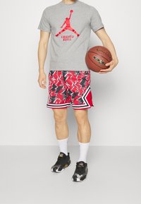 Camiseta gris con gráfico rojo de los Chicago Bulls, pantalones cortos de patrón zigzagueante rojo y negro, calcetines blancos y zapatillas deportivas negras, sosteniendo un balón de baloncesto.