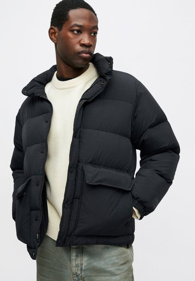 Vyras, vilkintis juodą pūkinę striukę ant kreminės spalvos megztinio ir šviesiai mėlynų džinsų, žiūri į šoną, rankas įsidėjęs į striukės kišenes.