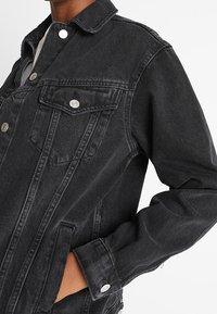 Svart denimjacka med silverfärgade metallknappar, bröstficka med lock och hand avslappnat placerad i sidfickan.