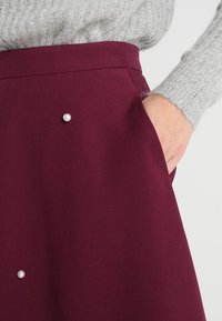 Burgunderfarbene Rock aus strukturiertem Stoff, mit zwei dekorativen Perlenakzenten an der Vorderseite und seitlichen Taschen.