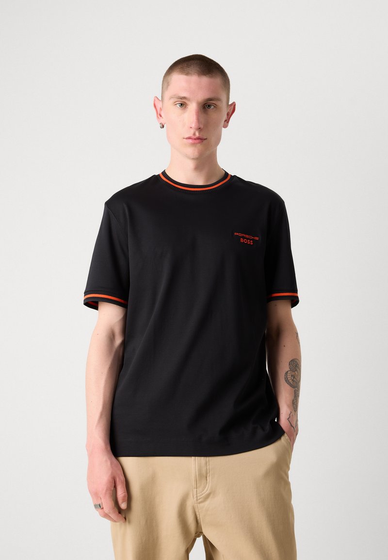 T-shirt noir à manches courtes avec des accents orange sur le col et les manches. Présente un petit logo en rouge sur le côté gauche de la poitrine. Tissu lisse.