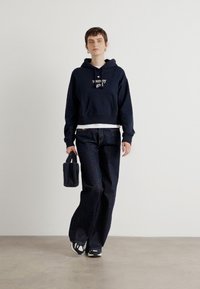 Cappuccio corto navy con il logo "tommy girl", abbinato a pantaloni a gamba larga in denim scuro. La modella tiene in mano una piccola borsa nera e indossa sneaker.