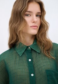 Camicia verde con texture, con bottoni e righe verticali, colletto e taschino. Presenta un materiale leggero e trasparente e bottoni bianchi.