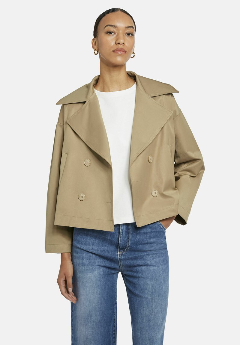 Vrouw in een beige cropped dubbelknopig jack, witte blouse, blauwe jeans en gouden creolen op een effen achtergrond.