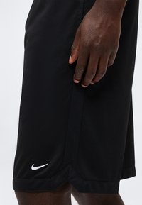 Main posée à côté d'un short de sport noir Nike avec le logo blanc Swoosh près de l'ourlet sur un fond clair.