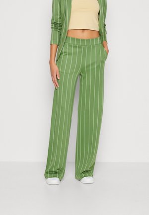 Kvinde iført grønne, stribede vidde-ben bukser og matchende blazer, beige crop top, hvide sneakers, stående med hænderne i lommerne.