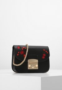 Musta nahkalaukku, jossa on punainen kukkakirjonta, kultaketju ja kultaklipsi, asetettu valkoiselle pedestalille yksinkertaista taustaa vasten.