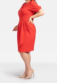 Červené šaty s krátkými rukávy, přiléhavým tělem a plisovanou sukní. Hladká textura, délka po kolena, kombinováno s nahými vysokými podpatky.