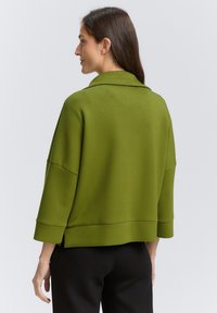 Femme aux cheveux bruns longs portant une veste verte olive ample et un pantalon noir, vue de dos sur un fond gris clair uni.