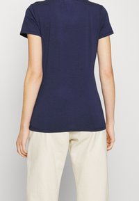 Person klädd i en åtsittande marinblå kortärmad t-shirt och lösa krämfärgade byxor, stående med armarna avslappnat vid sidorna.