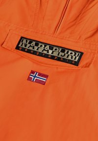 Giacca arancione con un evidente patch del logo "NAPAPIJRI geographic" in nero e grigio e un piccolo dettaglio del flag norvegese ricamato.