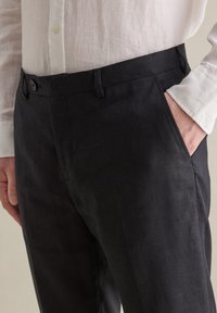 Pantalones de lino negros con un corte ajustado, que incluyen bolsillos laterales y una cinturilla con botón. La tela tiene un aspecto texturizado y un acabado mate.
