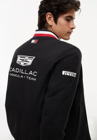 Άνδρας που φοράει μαύρο μπουφάν της ομάδας Cadillac Formula 1 με γιακά Tommy Hilfiger και λογότυπο Pirelli στο μανίκι, κοιτάζοντας πλάγια.