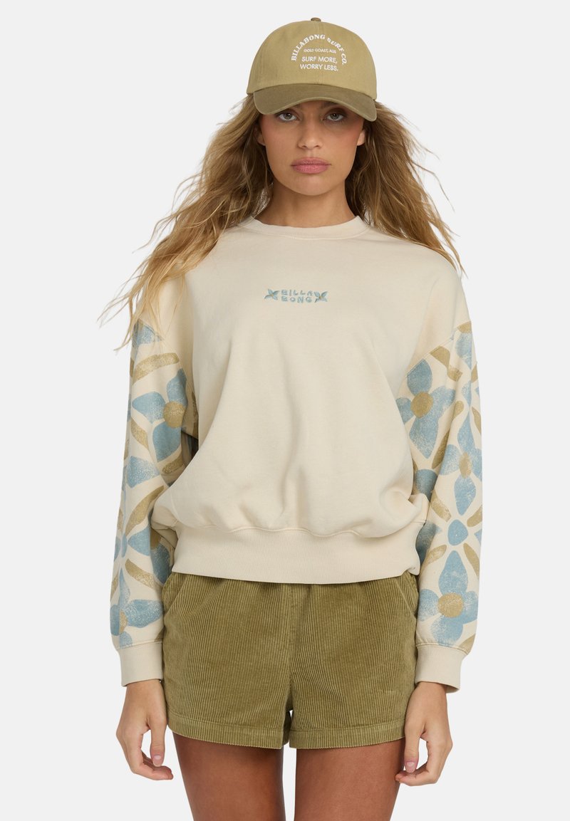 Sudadera crema con mangas de estampado floral en azul y dorado; cuenta con puños y cintura acanalados. Pantalones cortos de pana verde oliva. Gorra de color caqui claro.