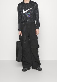 Personne portant un t-shirt à manches longues noir à motifs Nike avec le numéro 72, un pantalon cargo noir, des baskets noires, tenant un sac fourre-tout noir.