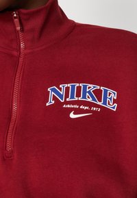 Vinröd Nike zip-up sweatshirt med hög krage. Har vita och blå logotyper: "NIKE" och "Athletic dept. 1972." Mjuk textur, avslappnad design.