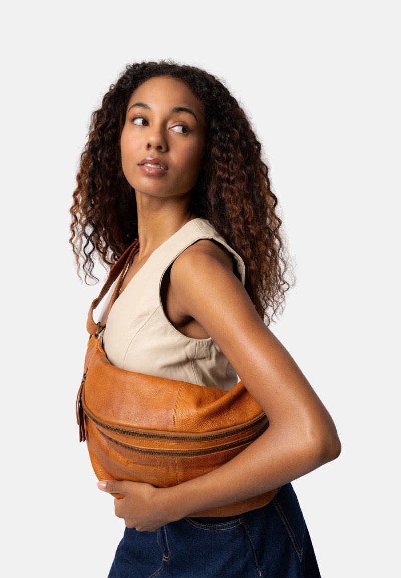 Bruine leren schoudertas met een ritssluiting, gebogen vorm en een gestructureerde afwerking, gehouden tegen een neutrale achtergrond.