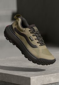 Beige and olive green sneakers with textured upper, black sole, prominent laces, and branding details. Accented with a red heel tab.