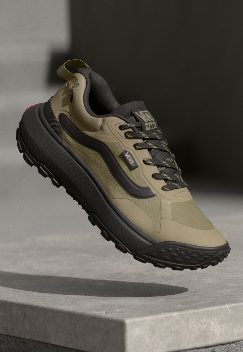 Beige and olive green sneakers with textured upper, black sole, prominent laces, and branding details. Accented with a red heel tab.