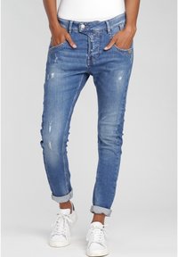 Persoon in opgerolde slim-fit blauwe versleten jeans en witte sneakers, met de handen in de voorzakken, staand tegen een grijze achtergrond.