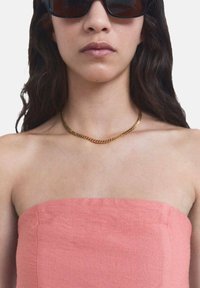 Top in lino rosa senza spalline con una texture liscia, accompagnato da una collana di catena dorata e occhiali da sole rettangolari scuri.