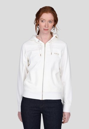 Haut à capuche zippé blanc avec un cordon de serrage et des accents dorés. Design simple avec des poignets et un ourlet côtelés, associé à un jean foncé.