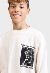 Cream-colored long-sleeve shirt made of cotton, featuring a black graphic print with text on the front. Round neckline and relaxed fit.