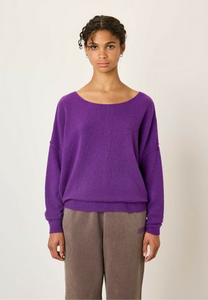 Lilla strikket trøje med bred, dyb halsudskæring og dropped shoulders. Blød tekstur. Sammen med brune, løstsiddende bukser.