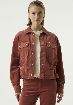 Mujer con el pelo corto castaño claro lleva una chaqueta de pana color óxido y pantalones a juego sobre un jersey claro, de pie frente a un fondo liso.