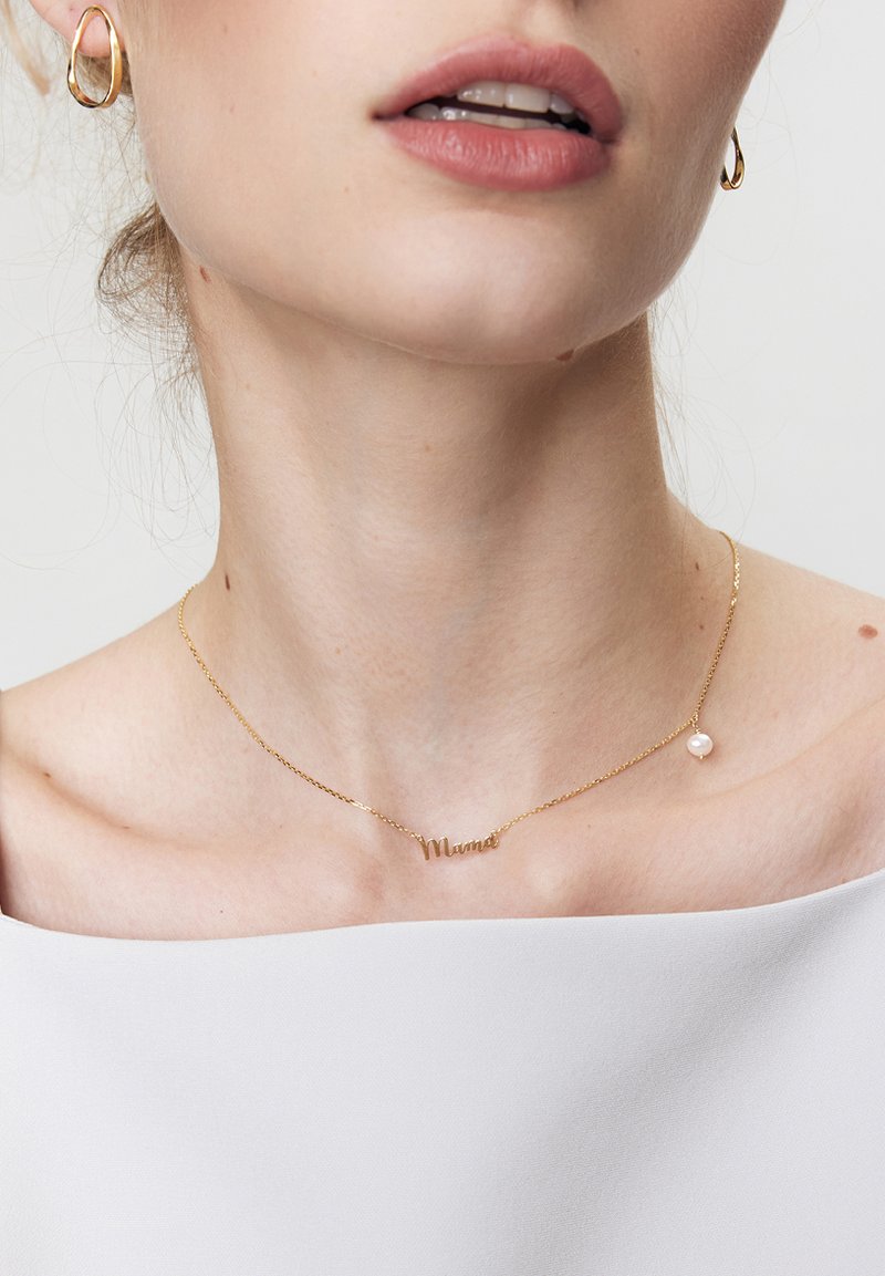 Złoty naszyjnik z napisem "Mama", z małym perłowym akcentem. Noszony na gołej skórze, w zestawieniu z białą bluzką bez ramiączek.